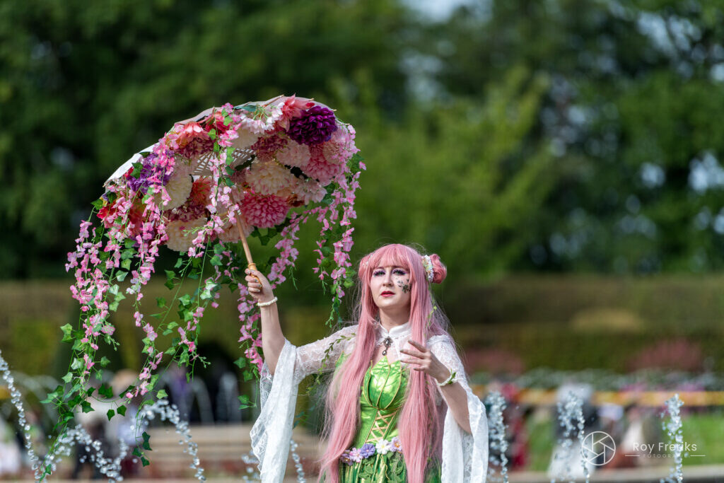 Vrouw met roze haar onder een paraplu vol bloemen in de kasteeltuin